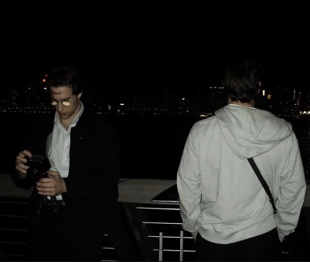 Two people at the railing by the water, with the Hong Kong skyline at night.